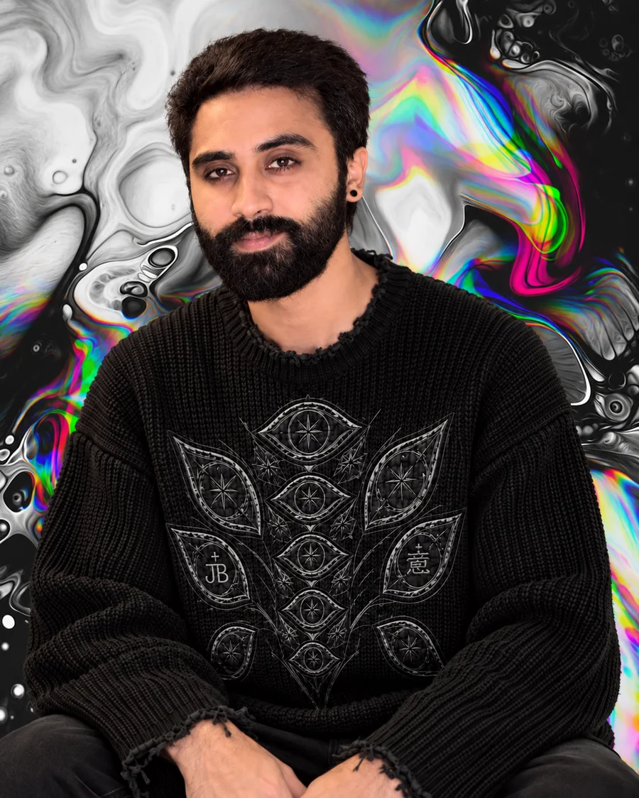 Vismay Hegde, founder of Meow Creative Haus, in a black knit sweater with embroidered eye and paisley motifs, against a black and white liquid backdrop with iridescent colour